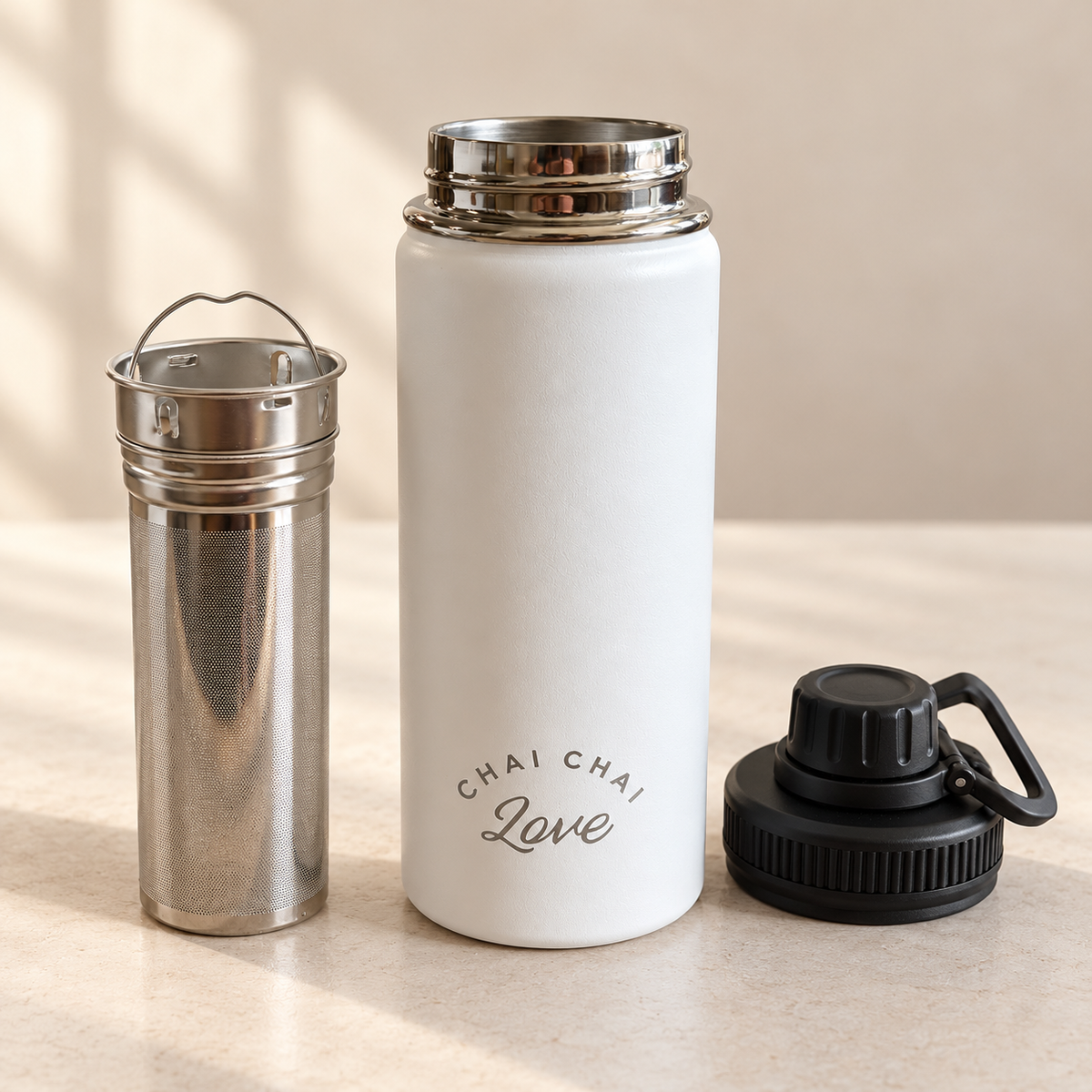 White insulated chai bottle beside its extra-long stainless steel tea infuser and black lid